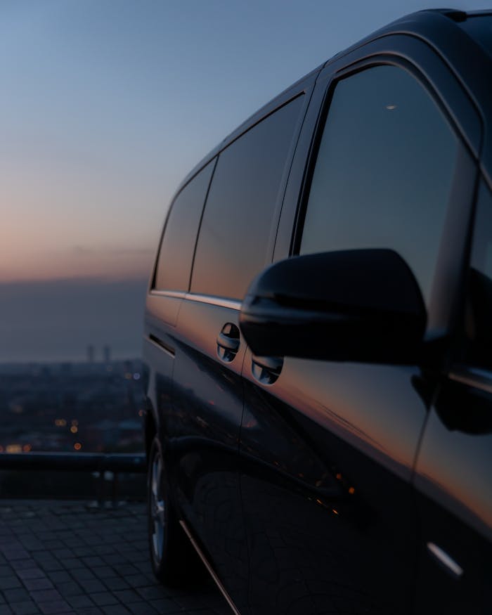 A luxury black car with tinted windows parked at a city overlook during dusk, reflecting twilight hues.