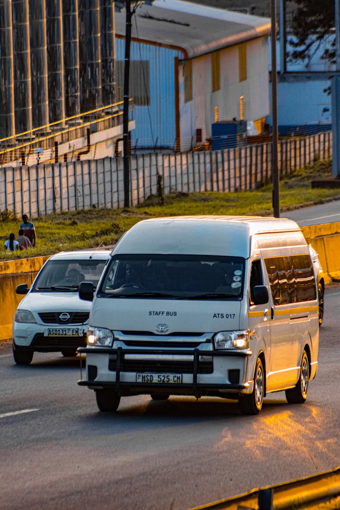A white minibus and car drive along a road in Matsapha, showcasing local transportation and infrastructure in daylight.